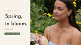 Spring Edit — 20% off. Woman in a blue floral dress in a flower garden wearing layered ANETT gold necklaces and hoop earrings.