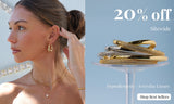 A split jewellery promotional image featuring a woman wearing gold hoop earrings, layered gold necklaces, and a gold bracelet on the left side, with sparkling crystal décor in the background. The right side shows a stack of gold and silver bangles displayed on a clear stand against a soft blue backdrop, alongside text promoting a 20% sitewide sale and hypoallergenic everyday luxury pieces.