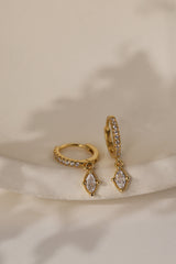 Pair of Sara gold marquise drop huggie earrings, angled on a cream dish