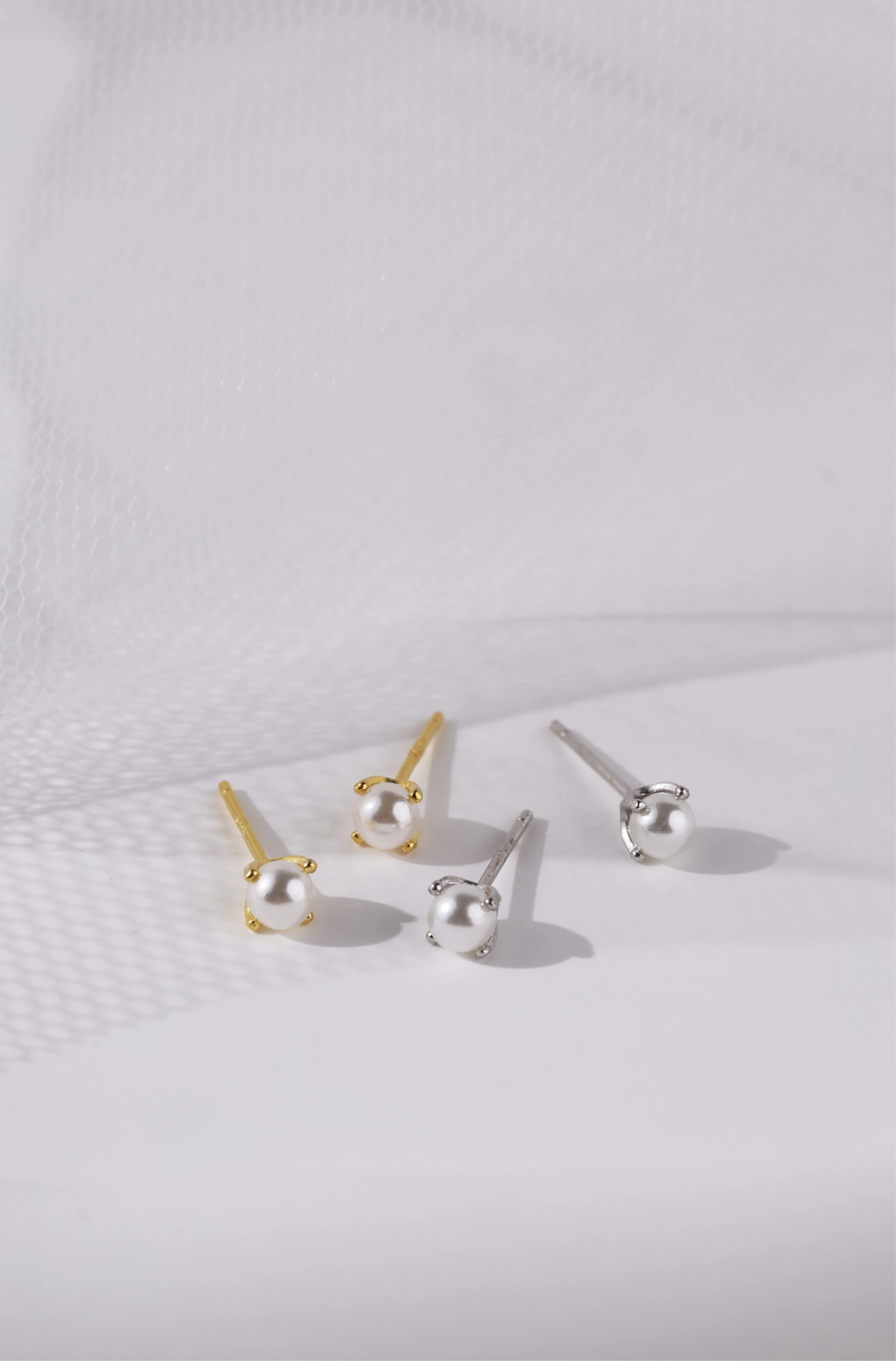 Four stud earrings arranged on a white textured surface, including two gold-toned earrings with white pearl-like beads and two silver-toned earrings with clear bead-like studs.