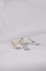 Four stud earrings arranged on a white textured surface, including two gold-toned earrings with white pearl-like beads and two silver-toned earrings with clear bead-like studs.
