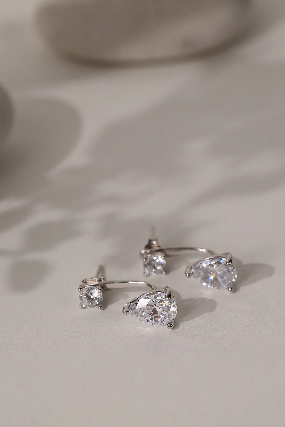 Sterling silver Adamas ear jacket earrings standing on cream linen, stud above a pear-cut drop.
