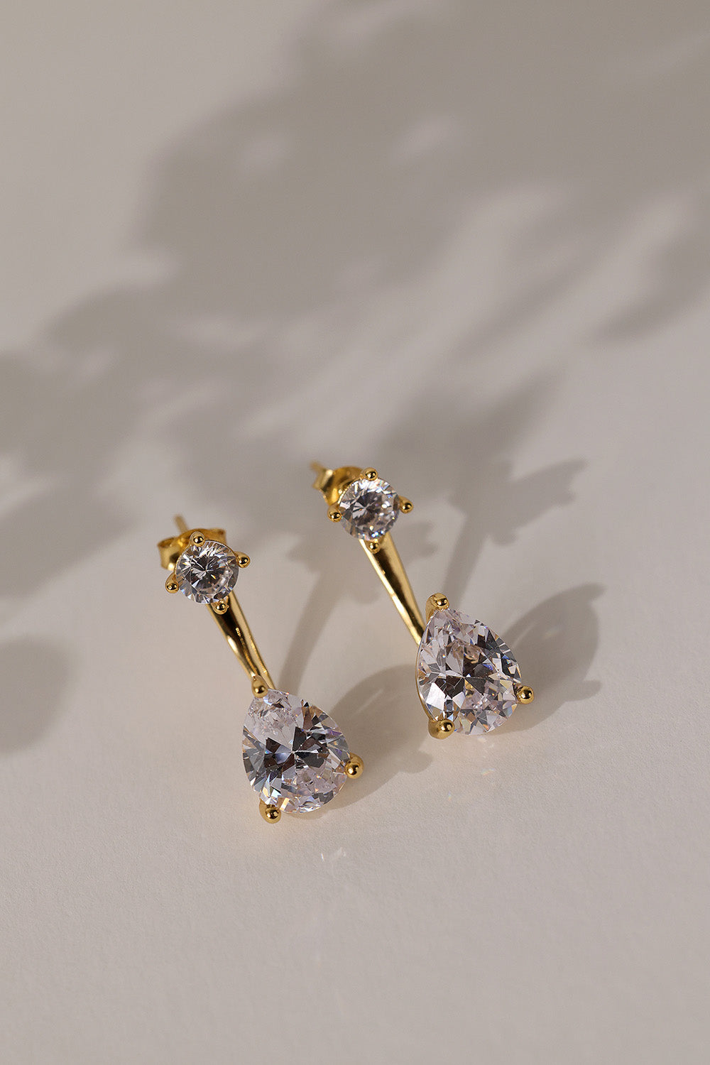 Gold Adamas ear jacket earrings lying flat on cream linen, stud above a pear-cut cubic zirconia drop.