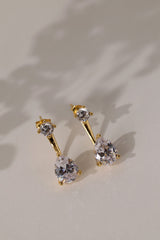 Gold Adamas ear jacket earrings lying flat on cream linen, stud above a pear-cut cubic zirconia drop.