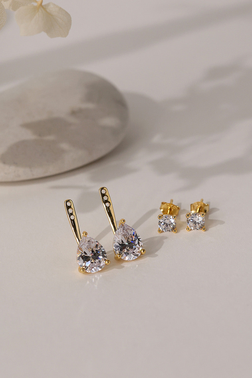 Gold Adamas ear jackets and round studs styled flat beside a stone dish and a dried flower.