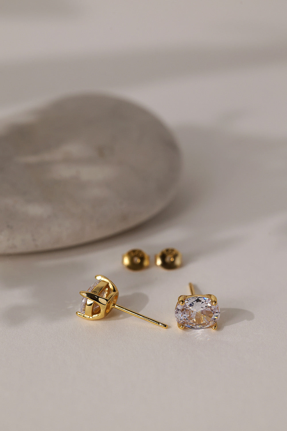 Gold Adamas oval stud earrings lying on cream beside a smooth stone, butterfly backs alongside.