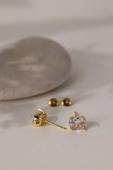 Gold Adamas oval stud earrings lying on cream beside a smooth stone, butterfly backs alongside.