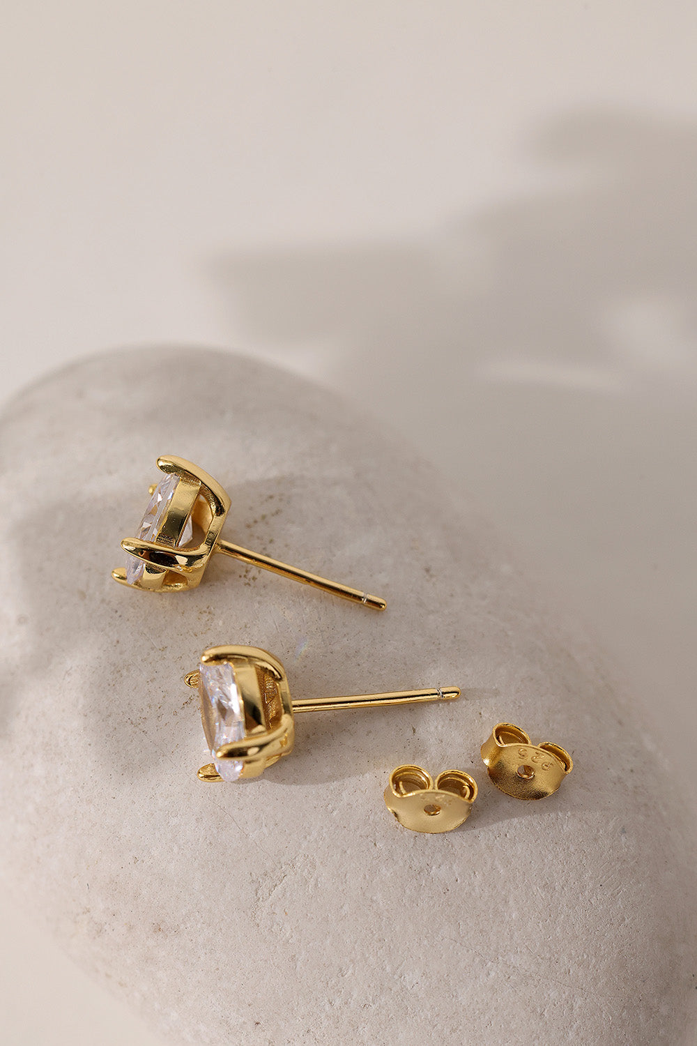 Pair of gold Adamas oval stud earrings resting on a pale pebble with their butterfly backs.