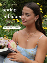 Spring Edit — 20% off. Woman holding flowers in a garden, wearing layered ANETT gold necklaces, hoops, rings and bracelets.