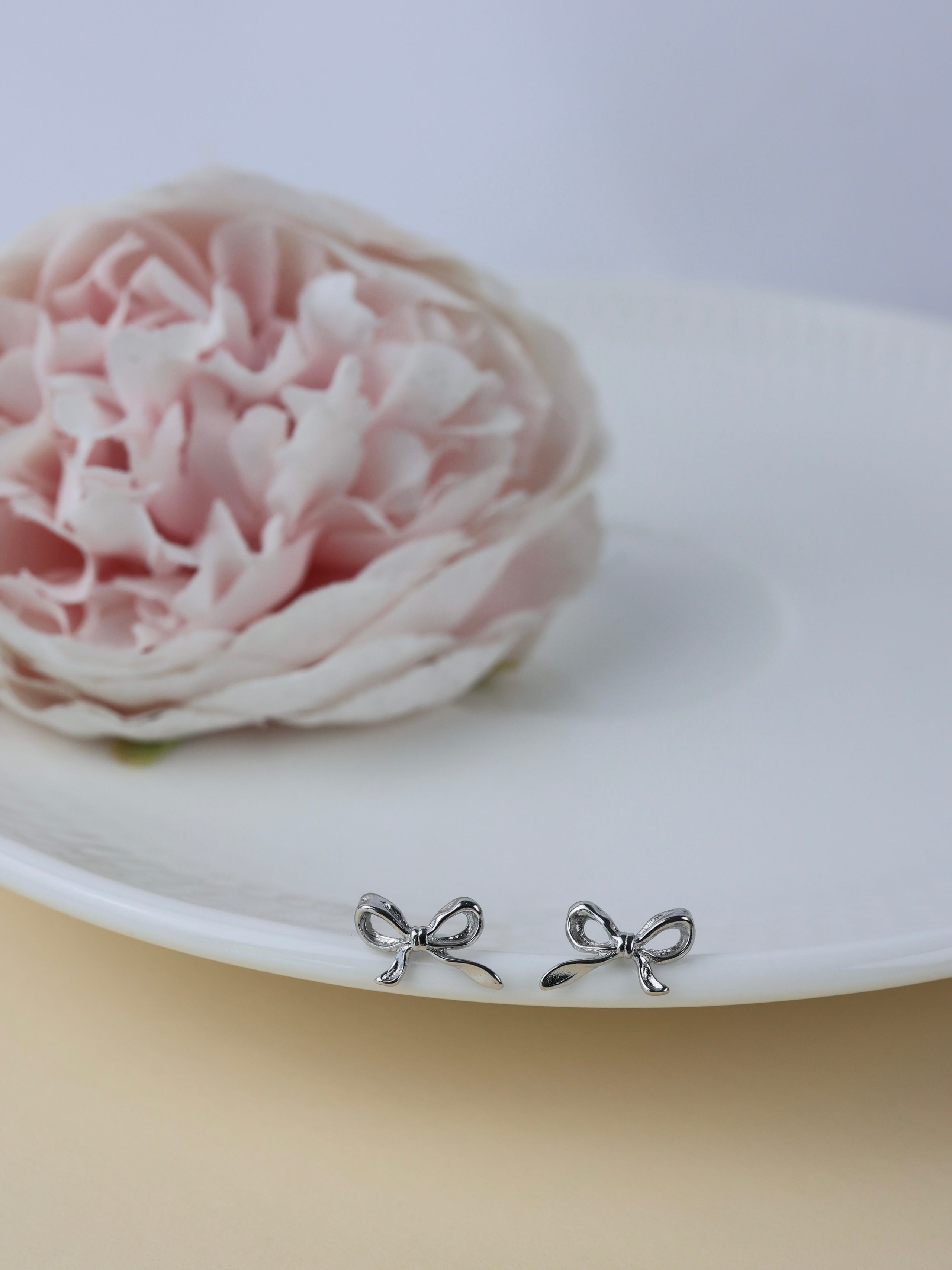 A pair of small silver bow stud earrings resting on the edge of a white plate, with a soft pink flower in the blurred background.
