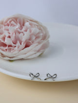 A pair of small silver bow stud earrings resting on the edge of a white plate, with a soft pink flower in the blurred background.