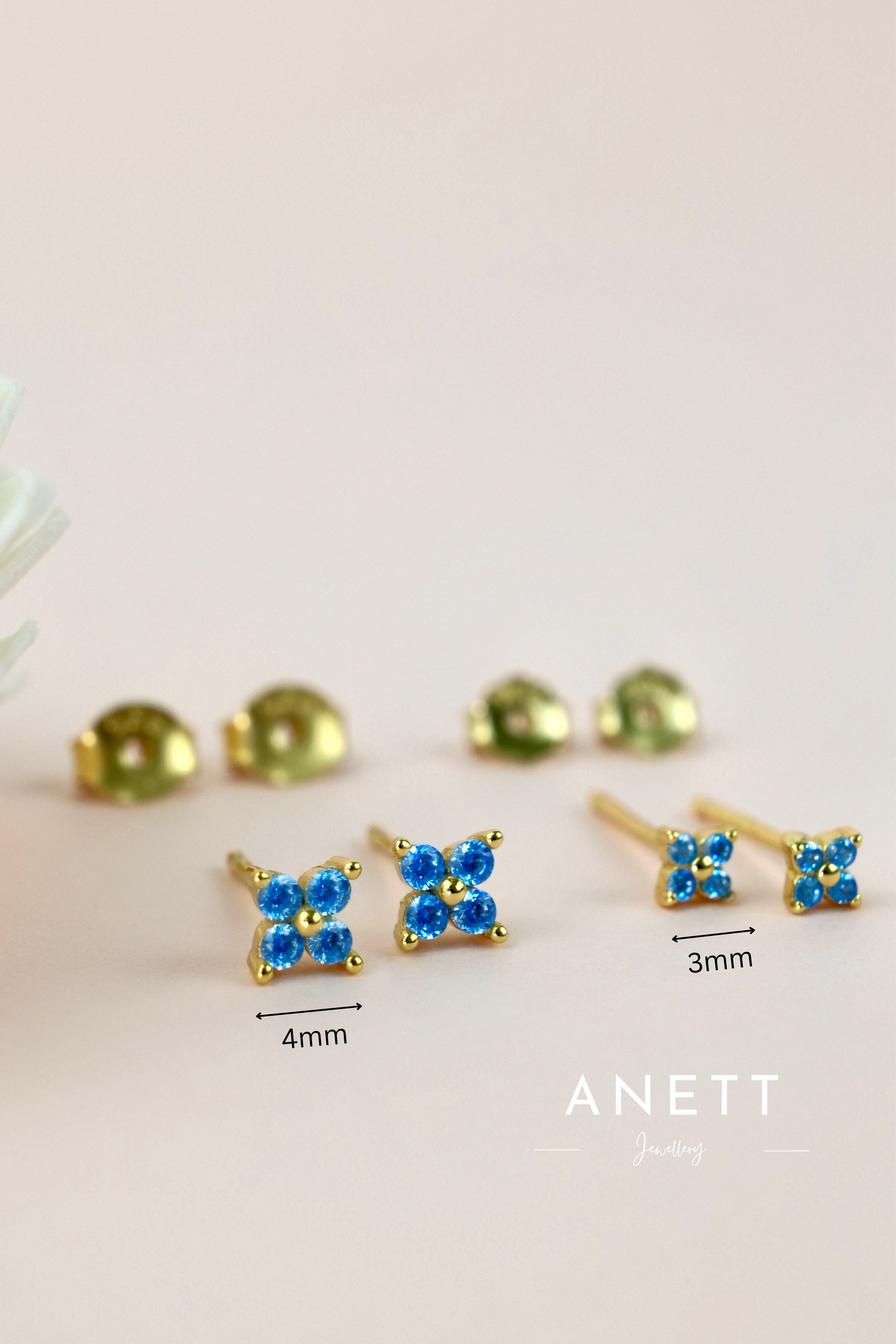 Gold-tone stud earrings with four clover blue cubic zirconia stones, shown in two sizes (4mm and 3mm) on a light beige background with brand logo 'ANETT Jewelry' at the bottom.