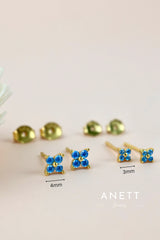 Gold-tone stud earrings with four clover blue cubic zirconia stones, shown in two sizes (4mm and 3mm) on a light beige background with brand logo 'ANETT Jewelry' at the bottom.