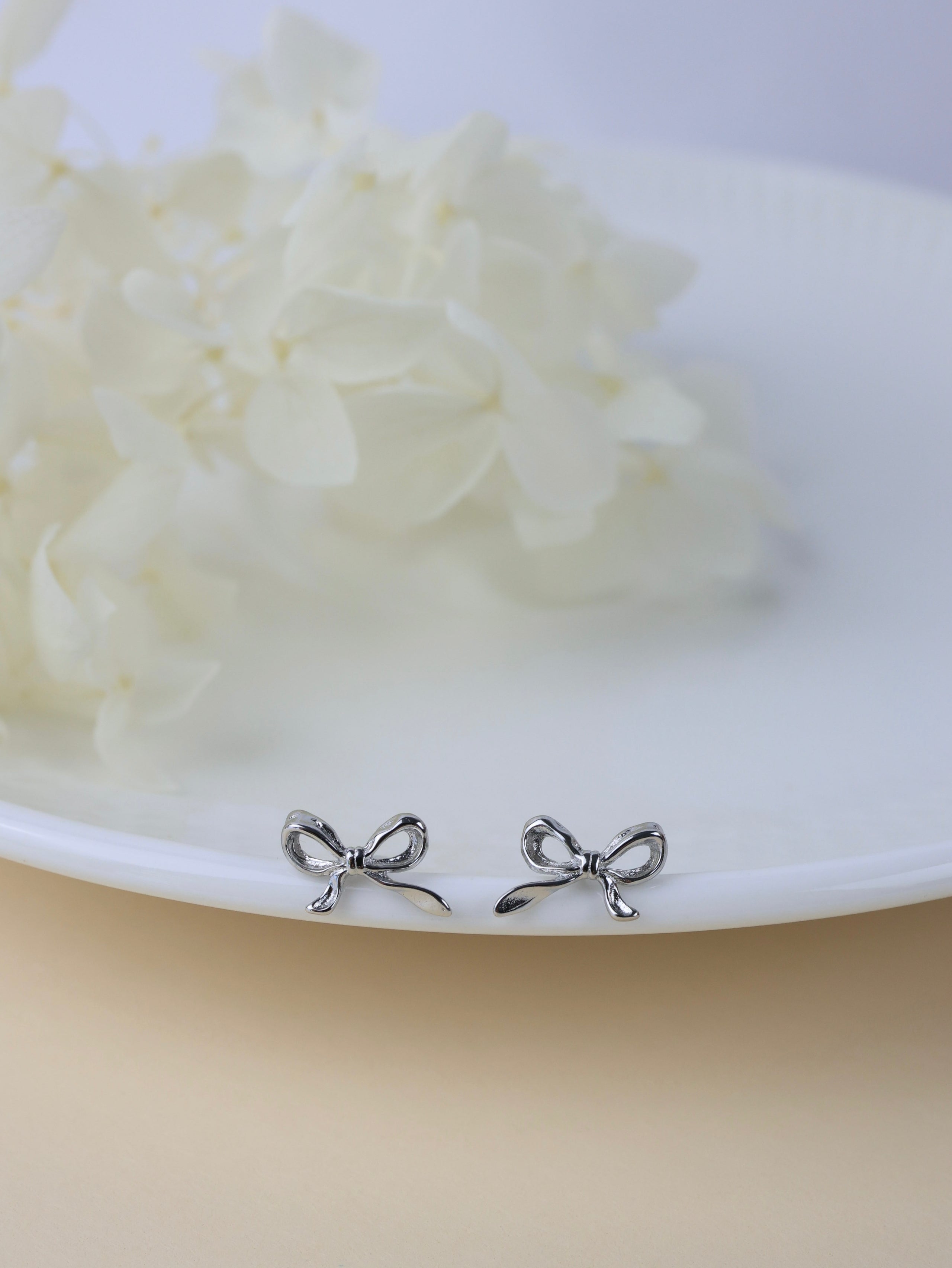 A pair of small silver bow stud earrings resting on the edge of a white plate with soft white flowers in the background.