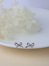 A pair of small silver bow stud earrings resting on the edge of a white plate with soft white flowers in the background.