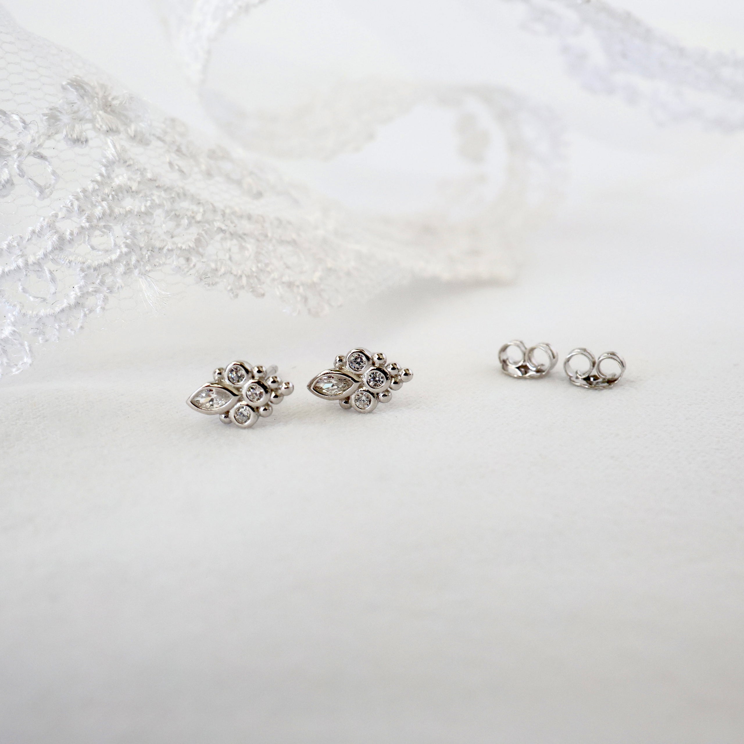 A pair of silver-toned floral stud earrings with clear rhinestones, displayed on a white fabric surface next to a pair of matching silver post backings, with white lace in the background.