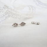 A pair of silver-toned floral stud earrings with clear rhinestones, displayed on a white fabric surface next to a pair of matching silver post backings, with white lace in the background.