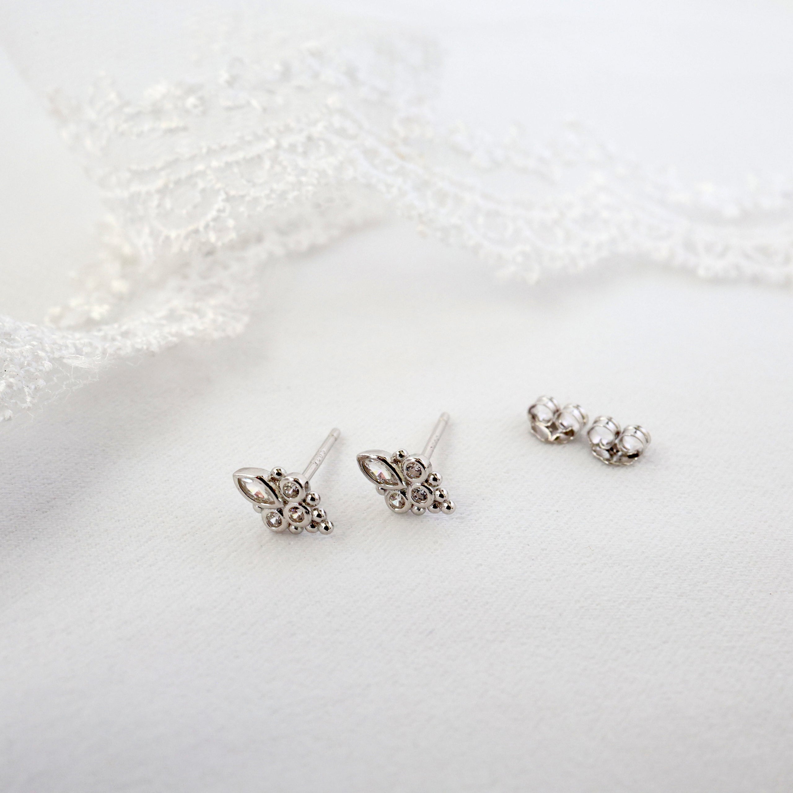 A pair of silver butterfly stud earrings with small clear rhinestones, displayed on a soft white textured surface with a white lace prop in the background.