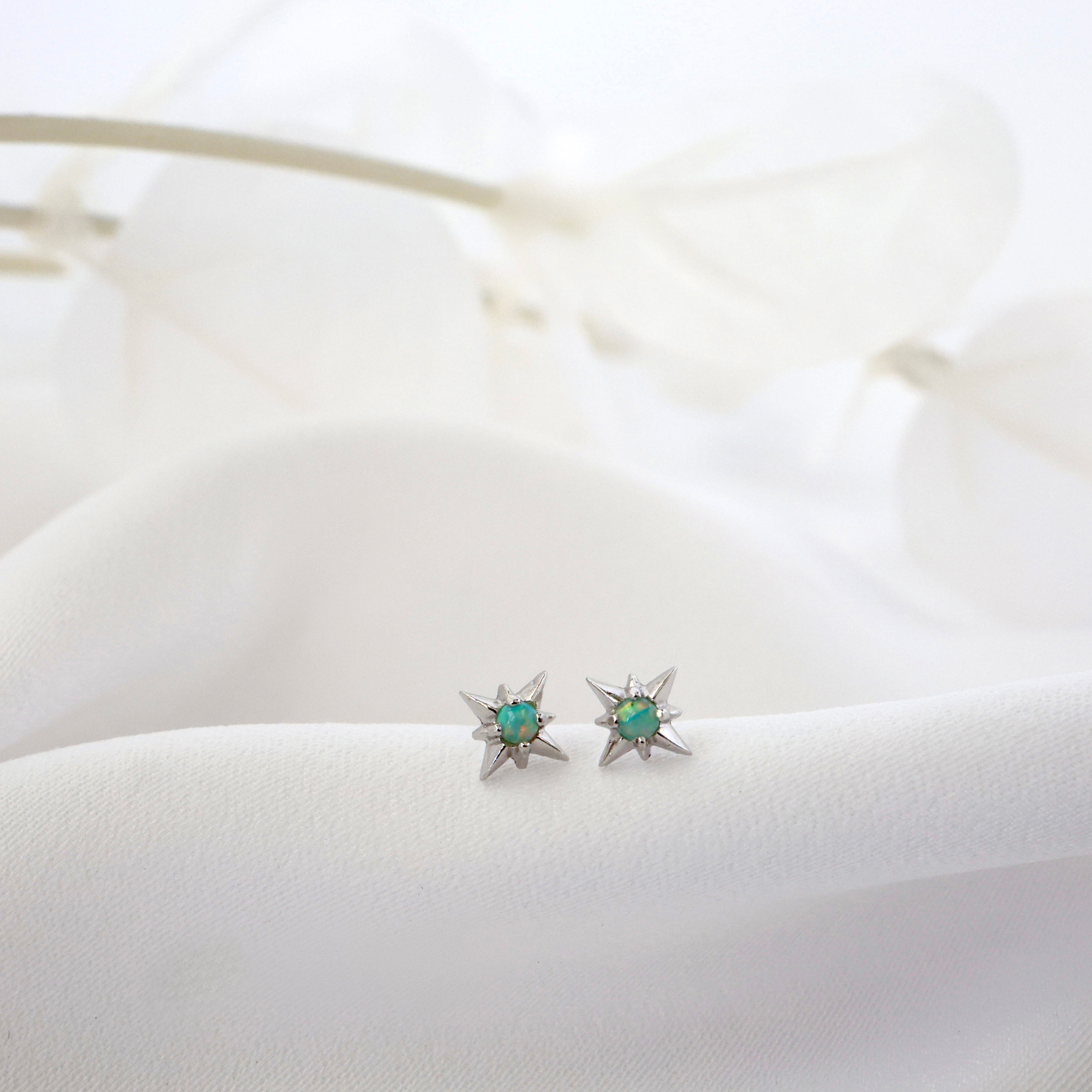 A pair of silver star-shaped stud earrings with round green opal  gemstones, resting on a soft white fabric surface.