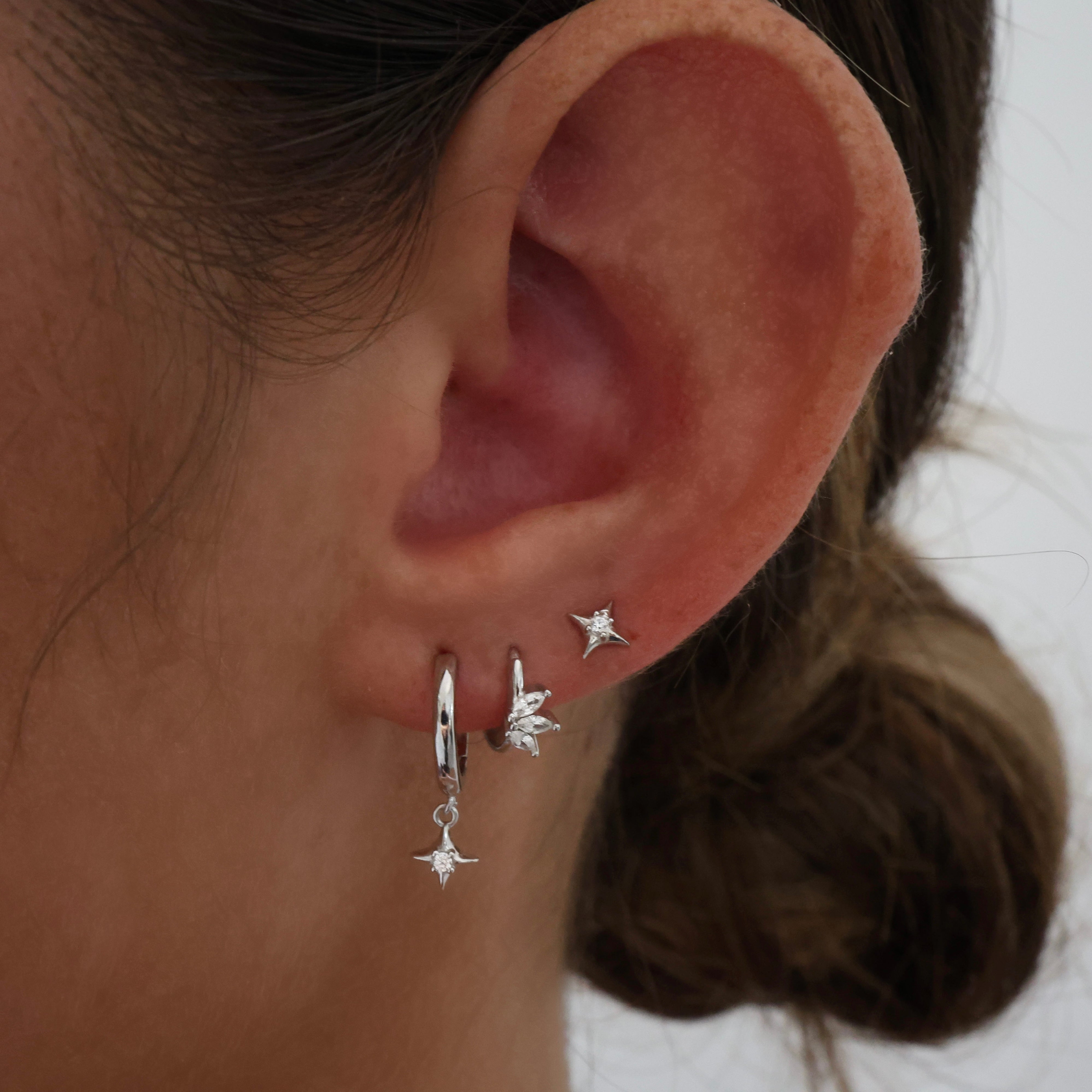 A close-up of a person's ear wearing three silver earrings: a hoop with a dangling star, a small star stud, and a leaf-shaped stud.