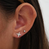 A close-up of a person's ear wearing two silver-toned stud earrings: a larger starburst earring with clear stones and a smaller matching star earring.