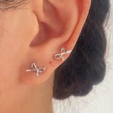 A close-up of a person's ear wearing two small silver bow-shaped stud earrings.