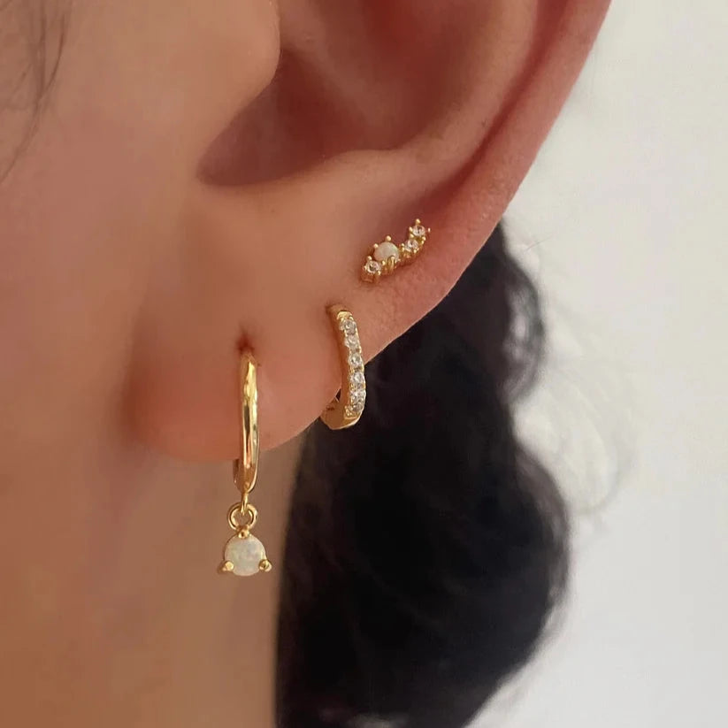 Close-up of a person's ear wearing three gold earrings: a small cluster stud, a huggie hoop with clear stones, and a drop hoop with a white teardrop-shaped stone.