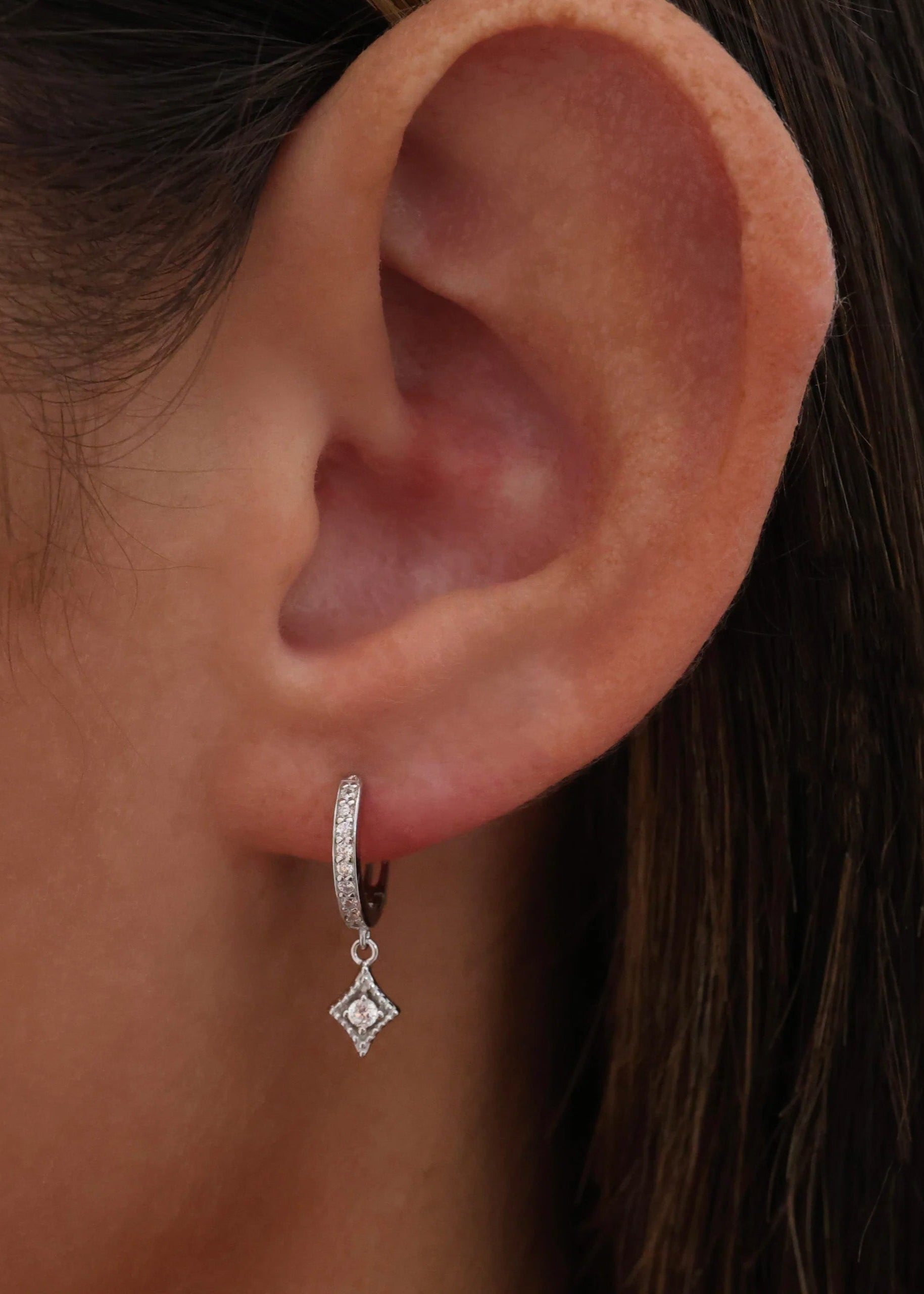 A close-up of a person's ear wearing a small silver huggie hoop earring with a dangling diamond-shaped charm set with clear stones.