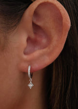 A close-up of a person's ear wearing a small silver huggie hoop earring with a dangling diamond-shaped charm set with clear stones.