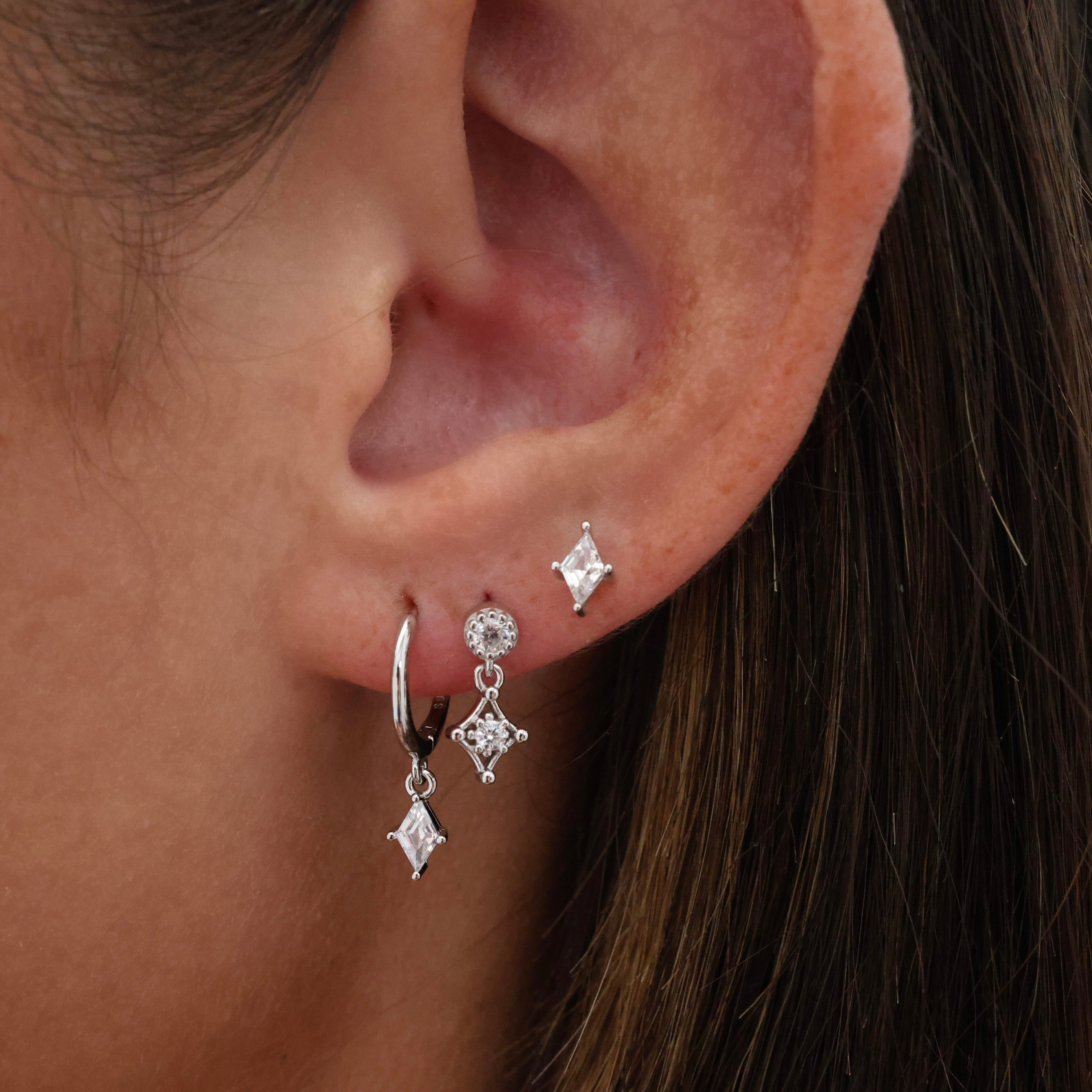 A close-up of a person's ear wearing silver hoop earrings with dangling clear gemstone diamond charms.