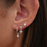 A close-up of a person's ear wearing silver hoop earrings with dangling clear gemstone diamond charms.