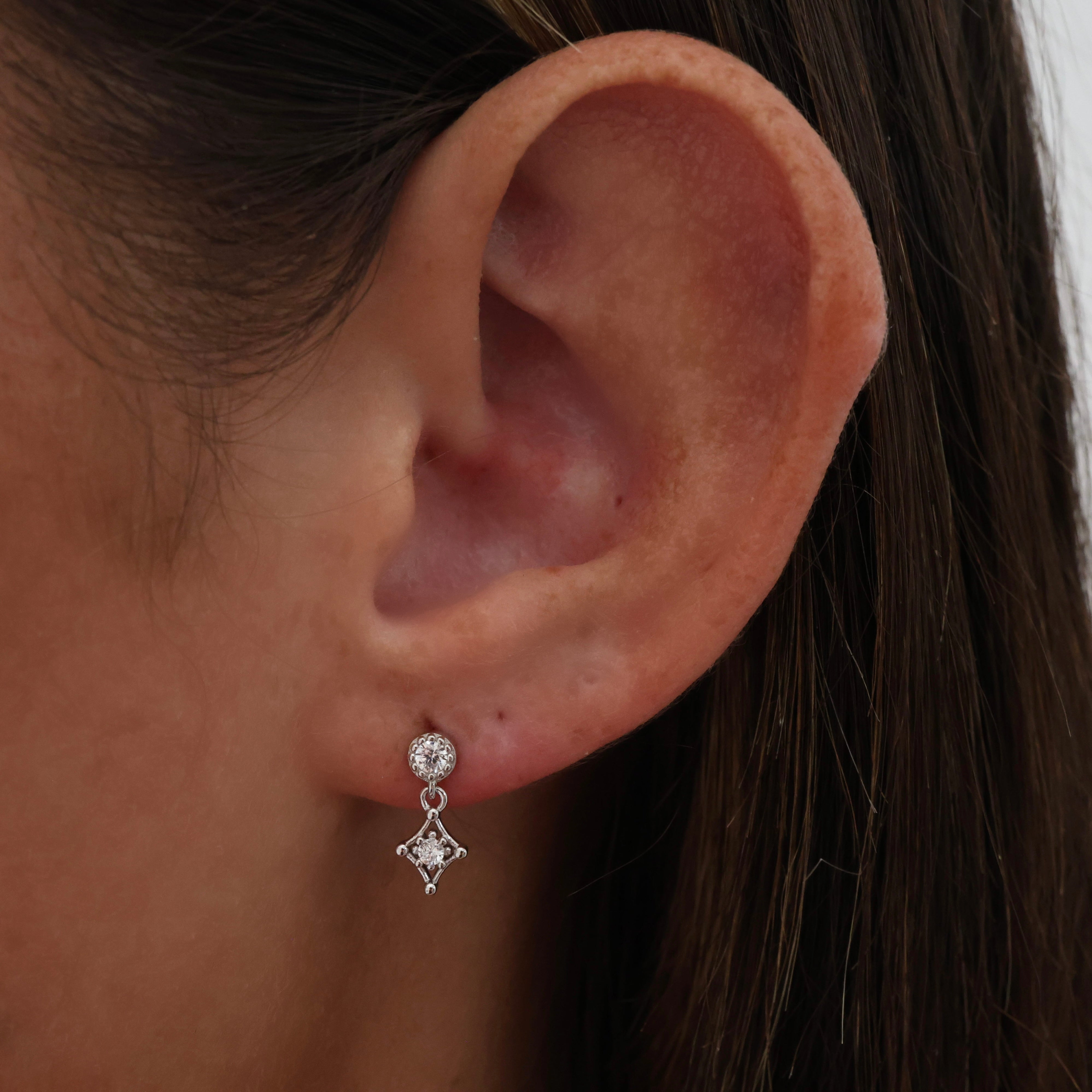 A close-up of a person's ear wearing a small silver dangle earring with clear gemstones.