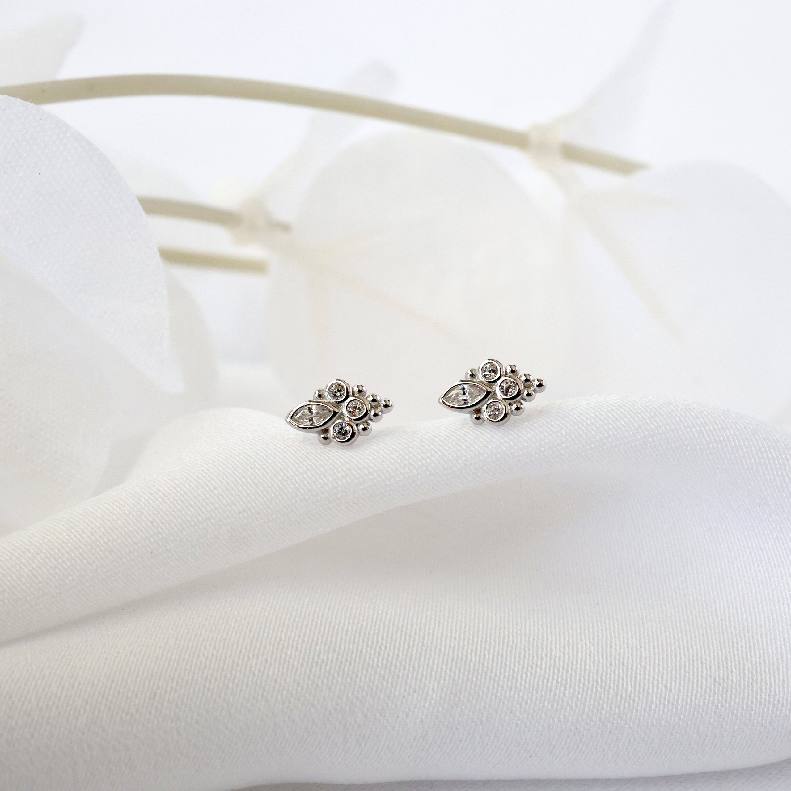 A pair of silver-toned leaf-shaped stud earrings resting on soft white fabric with blurred white flowers in the background.