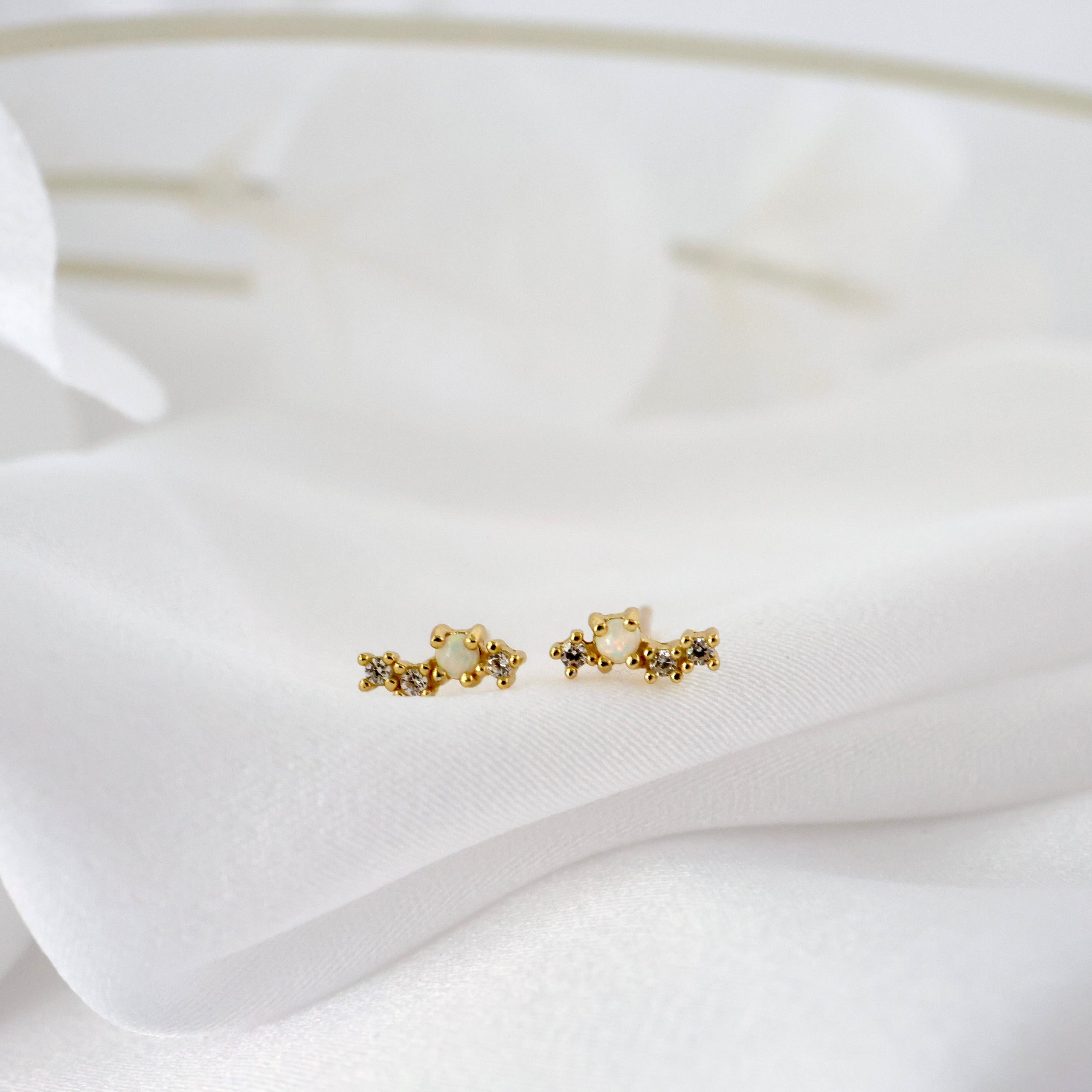 A pair of small gold stud earrings with a floral or starburst design, resting on a soft white fabric background.