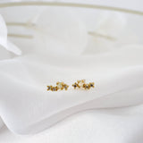 A pair of small gold stud earrings with a floral or starburst design, resting on a soft white fabric background.