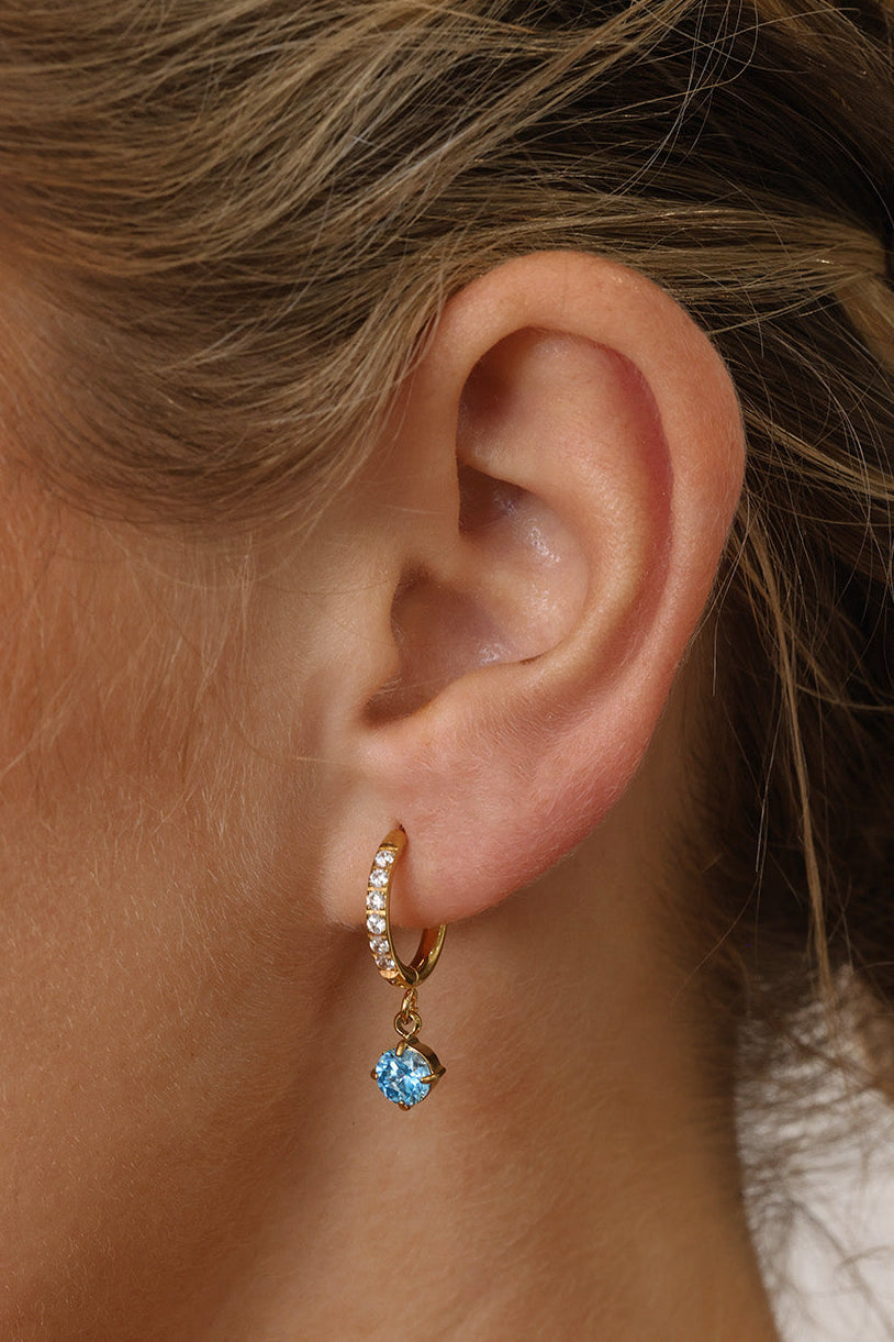 Gold pavé hoop earrings with clear stones and a dangling round blue birthstone, photographed on a soft cream background with blurred white florals.