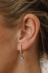 Gold pavé hoop earrings with clear stones and a dangling round blue birthstone, photographed on a soft cream background with blurred white florals.
