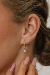 Gold pavé hoop earrings with clear stones and a dangling round blue birthstone, photographed on a soft cream background with blurred white florals.