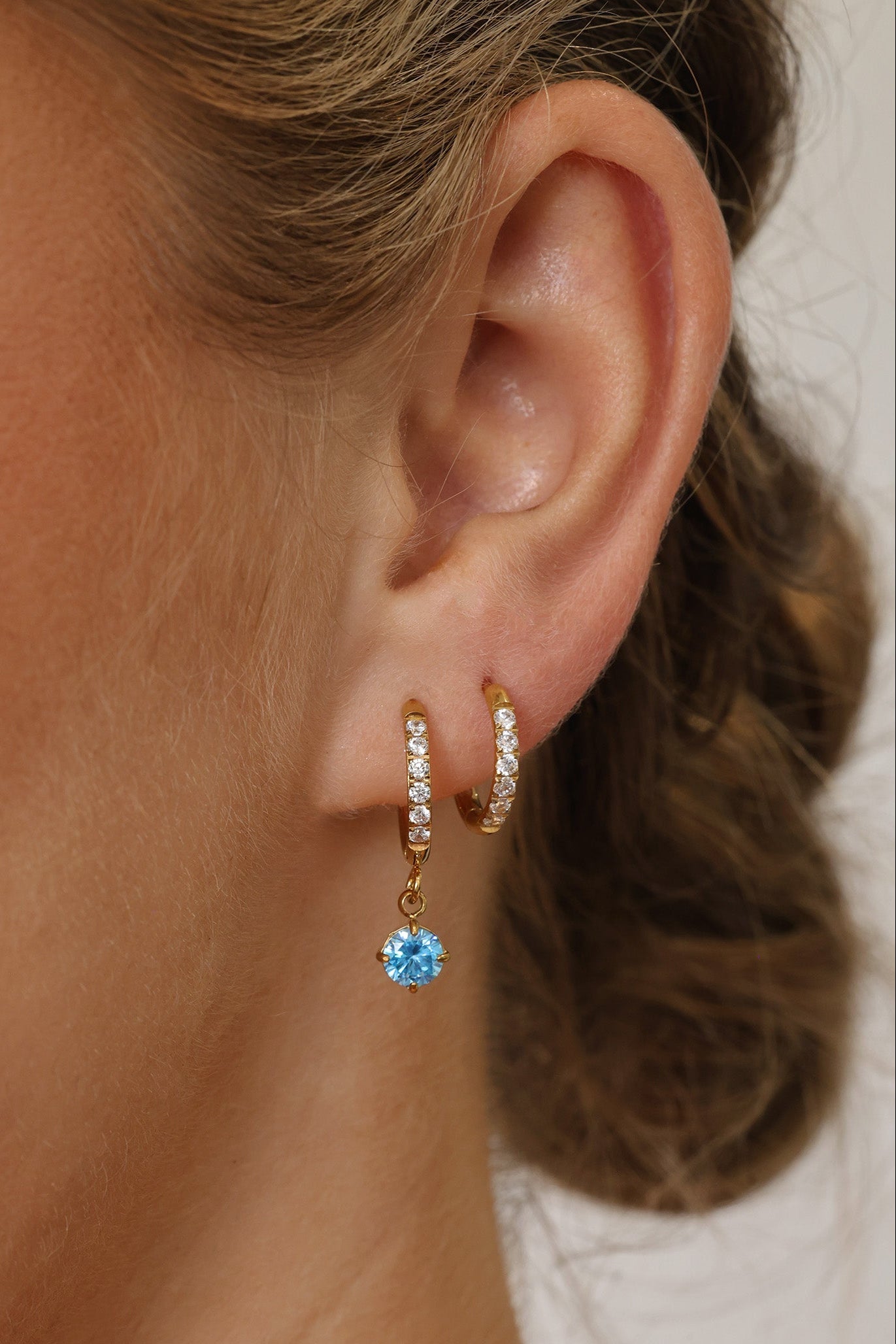 Gold pavé hoop earrings with clear stones and a dangling round blue birthstone, photographed on a soft cream background with blurred white florals.