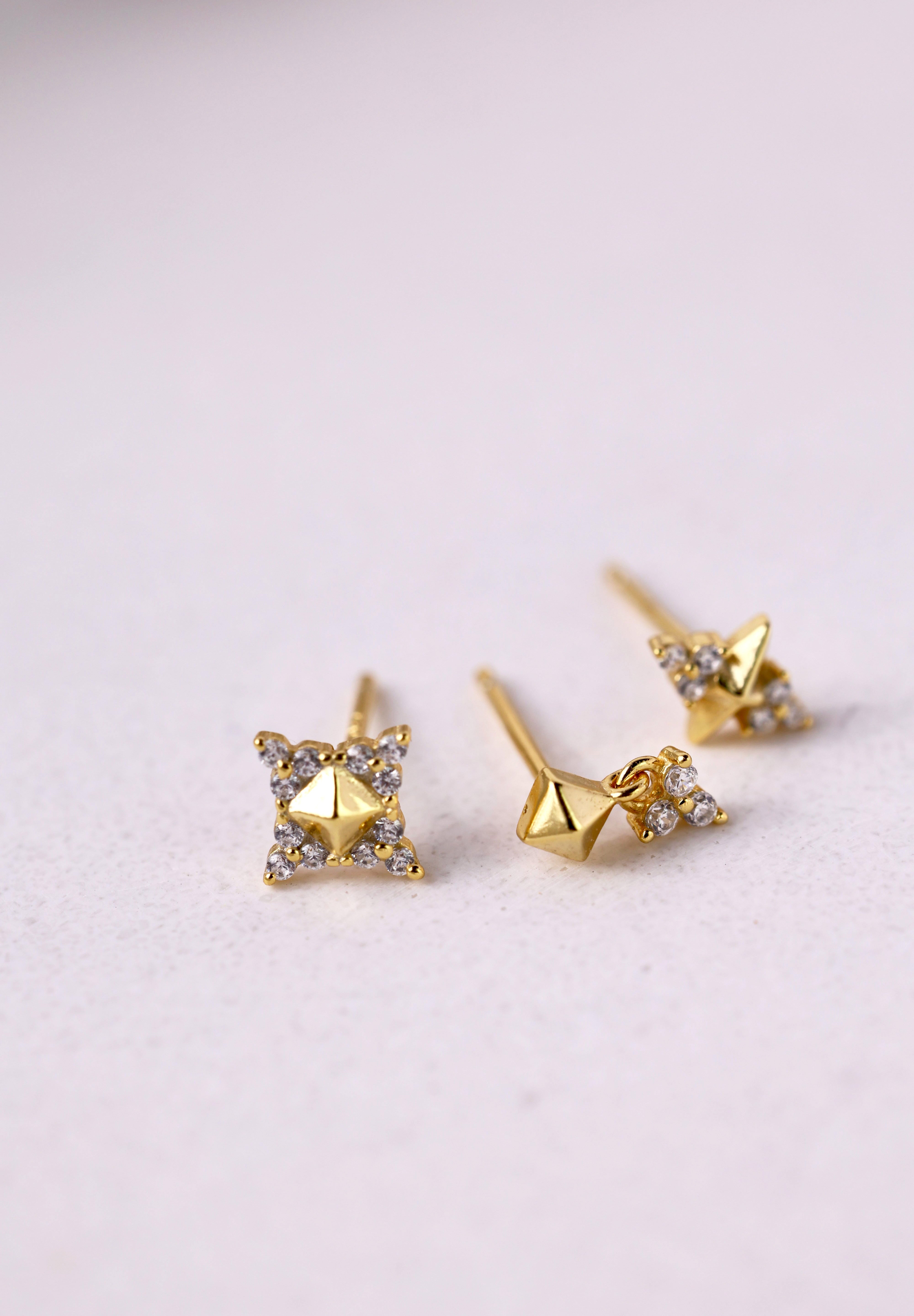 A pair of gold geometric stud earrings with small clear crystal accents, displayed on a light textured surface.