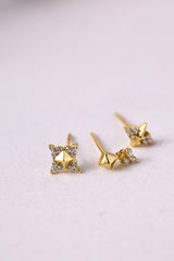 A pair of gold geometric stud earrings with small clear crystal accents, displayed on a light textured surface.