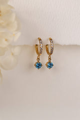 Gold pavé hoop earrings with clear stones and a dangling round blue birthstone, photographed on a soft cream background with blurred white florals.