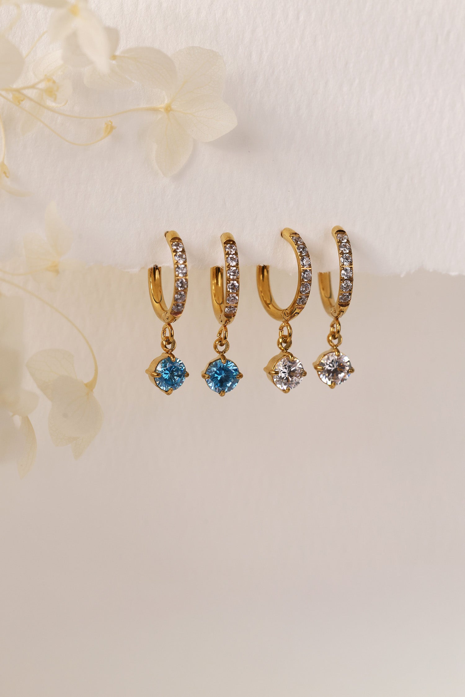 Gold pavé hoop earrings with clear stones and a dangling round blue birthstone, photographed on a soft cream background with blurred white florals.