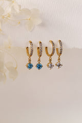 Gold pavé hoop earrings with clear stones and a dangling round blue birthstone, photographed on a soft cream background with blurred white florals.