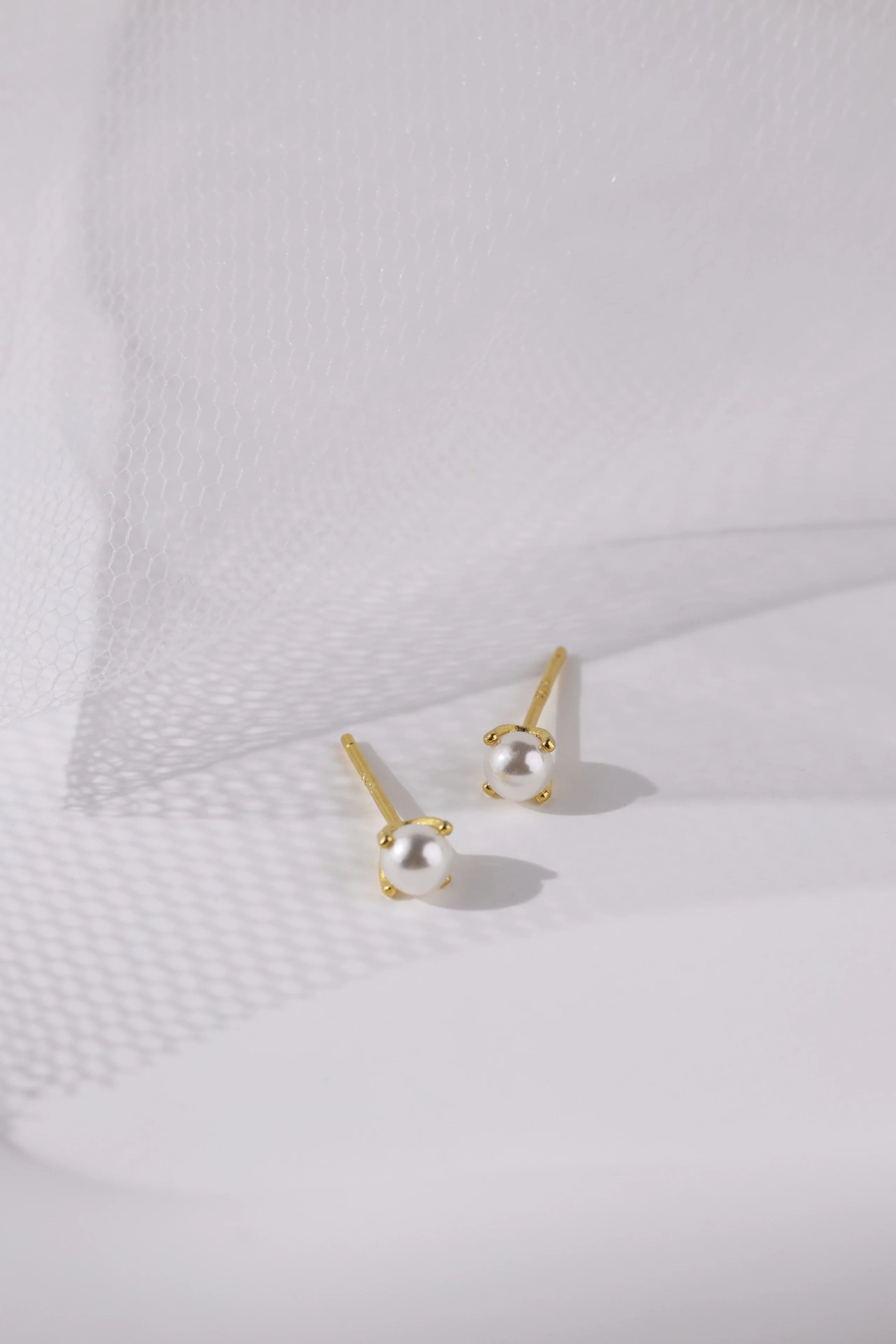 A pair of gold stud earrings with small white pearl-like beads, displayed on a clean white textured surface with soft shadows.