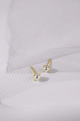 A pair of gold stud earrings with small white pearl-like beads, displayed on a clean white textured surface with soft shadows.