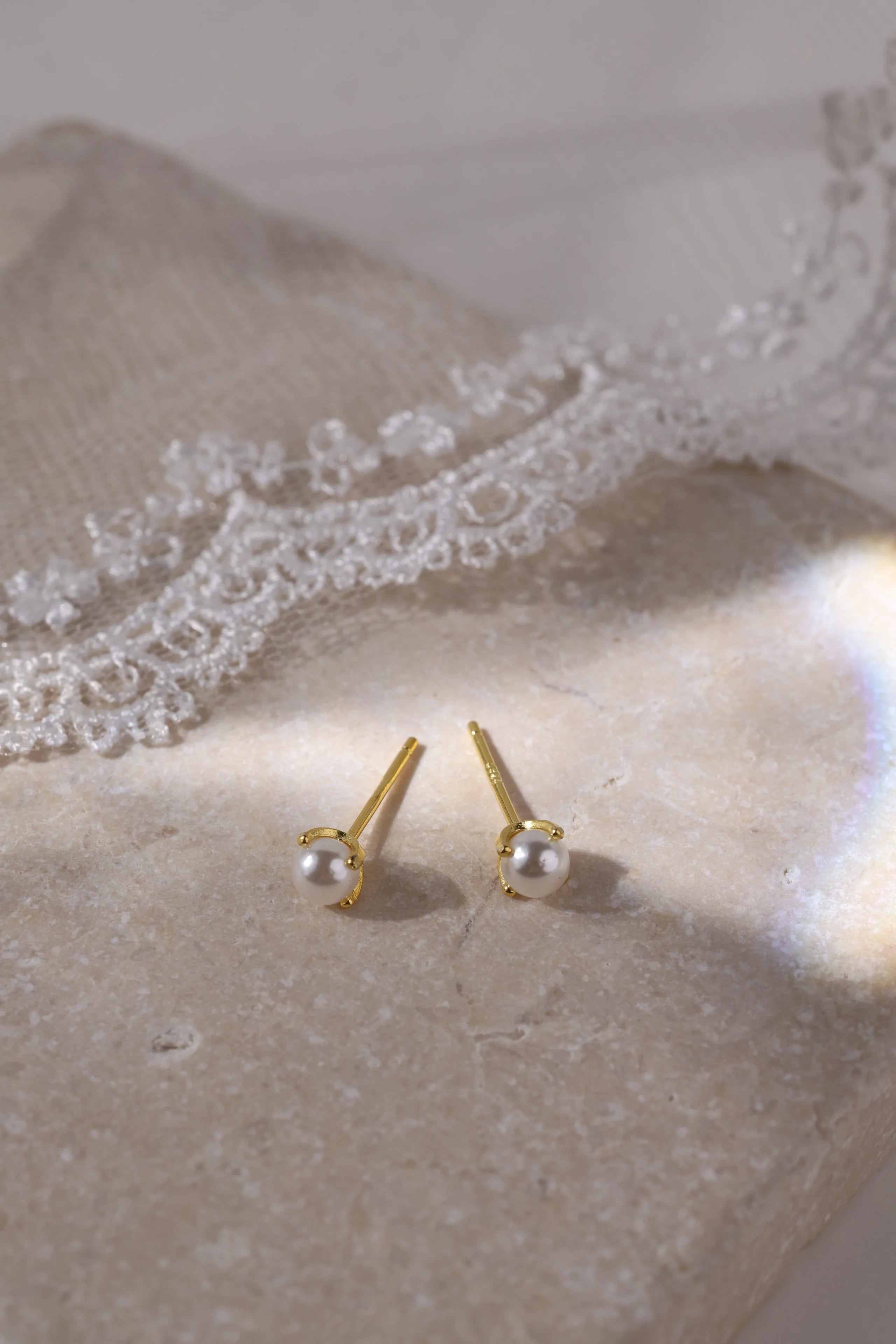 A pair of gold stud earrings with small white pearl-like beads, displayed on a textured beige surface with white lace in the background.