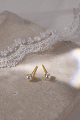 A pair of gold stud earrings with small white pearl-like beads, displayed on a textured beige surface with white lace in the background.