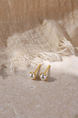 A pair of gold stud earrings with round white pearl-like beads, displayed on a textured beige surface with a frayed linen cloth in the background.