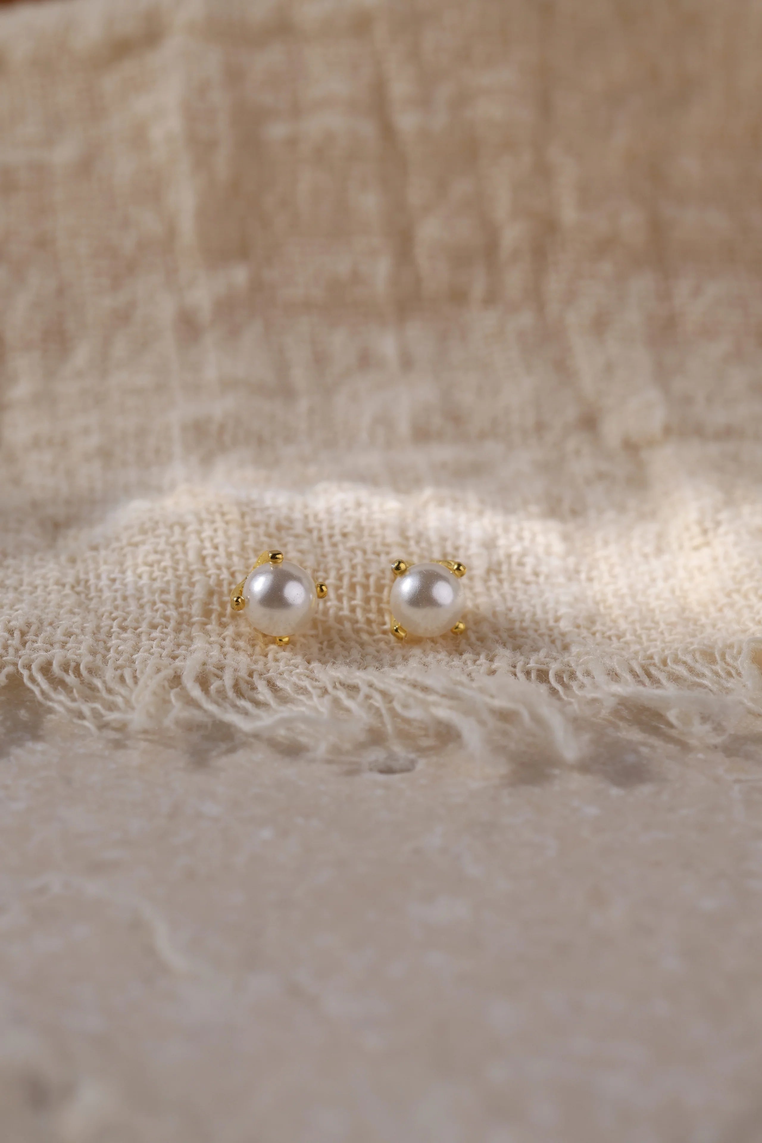 A pair of small gold stud earrings with white pearl-like beads, resting on a textured beige fabric background.